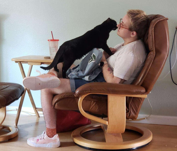 Tuesday, a white person, is seated in a brown leather chair. They're working on a sewing project while their black cat stands on their leg, standing nose to nose with Tuesday.
